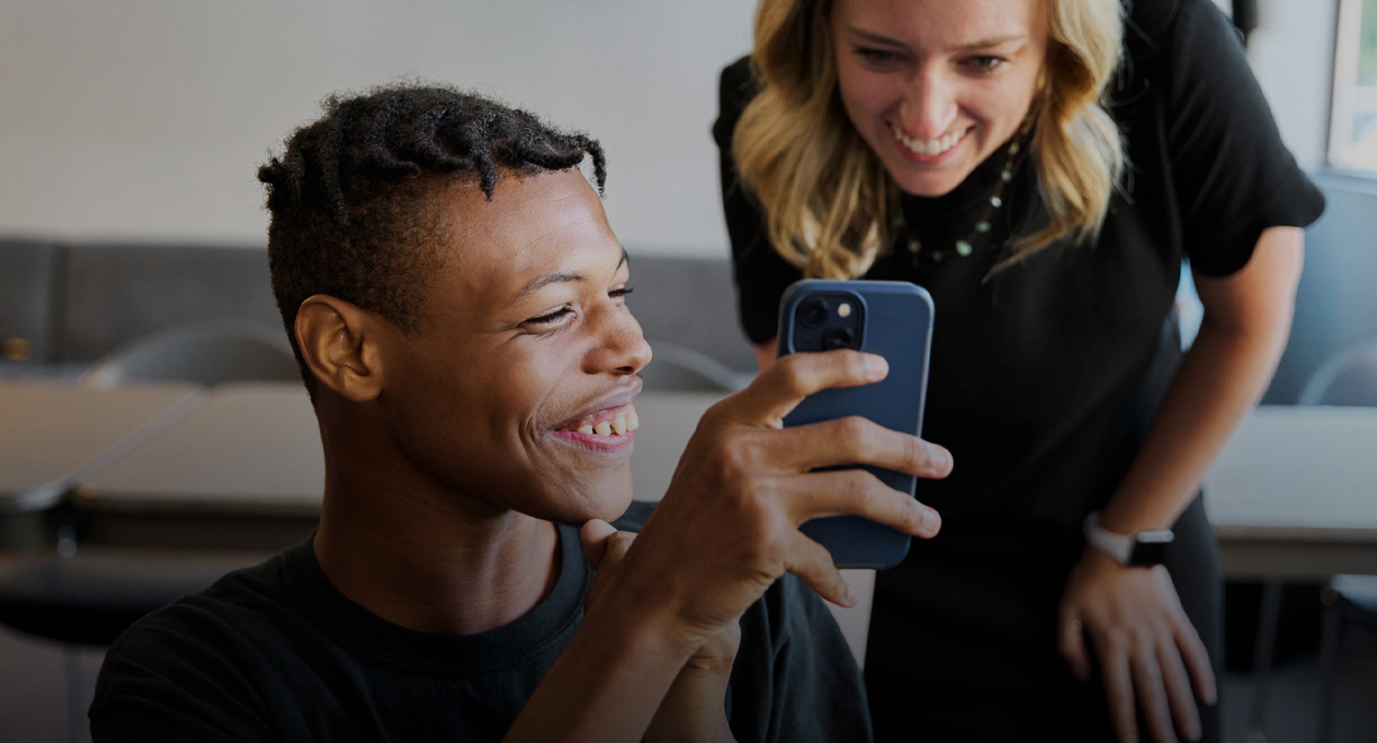 Foto von einem jungen Mann mit eingeschränktem Sehvermögen, der lächelt und einer lächelnden Frau, die hinter seiner linken Schulter steht, das Display seines iPhone zeigt