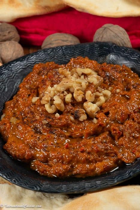 A bowl full of muhammara, a syrian red pepper sauce garnished with walnuts and surrounded by pita.